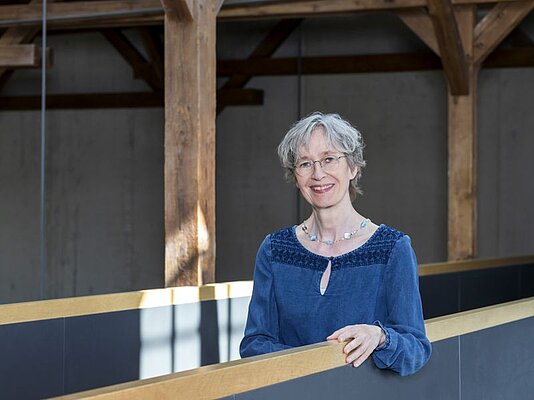 Eine Frau mit kurzen, grauen Haaren steht an einem Holzbalkon. Sie trägt ein blaues Oberteil mit einem dekorativen Ausschnitt und eine Kette. Im Hintergrund sind Holzstützen und eine schlichte Wand zu sehen. Die Frau lächelt freundlich in die Kamera.