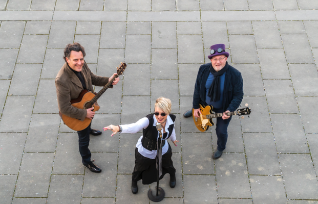 Drei Musiker stehen im Freien auf einem gepflasterten Boden. Eine Sängerin steht in der Mitte mit einem Mikrofon, während die beiden anderen Musiker je eine Gitarre halten. Alle blicken nach oben und wirken dynamisch und engagiert.