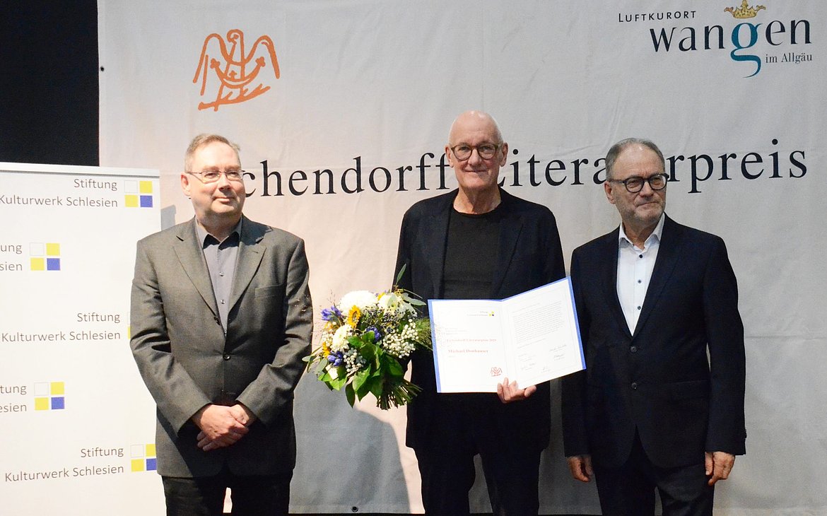 Drei Männer nebeneinander. Der Mittlere hat eine Urkunde und Blumen in der Hand.