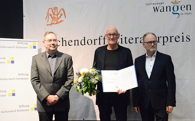 Drei Männer nebeneinander. Der Mittlere hat eine Urkunde und Blumen in der Hand.