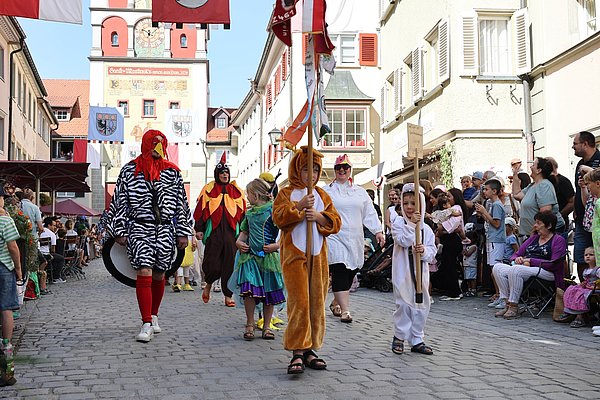Eine bunte Parade findet auf einer gepflasterten Straße statt. Menschen in Tierkostümen, darunter ein Hahn, ziehen vorbei. Kinder halten Schilder, während eine Menge Zuschauer an den Seiten steht. Flaggen und festliche Dekoration schmücken die Umgebung.
