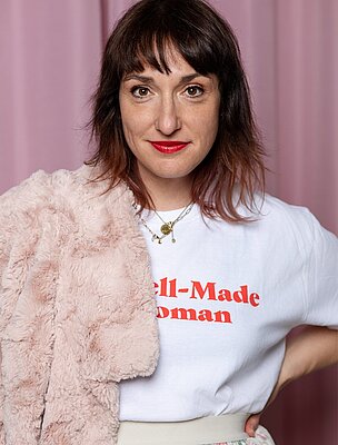 Eine Person mit mittellangen braunen Haaren trägt ein weißes T-Shirt mit der Aufschrift "Well-Made Woman" und hat eine rosa Jacke über der Schulter. Der Hintergrund ist in sanften Rosa- und Creme-Tönen gehalten. Die Person blickt direkt in die Kamera.