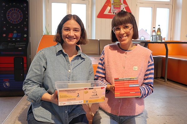 Zwei Frauen. Sie halten eine leere Box und eine symbolisch gebaute kleine Rampe in der Hand.