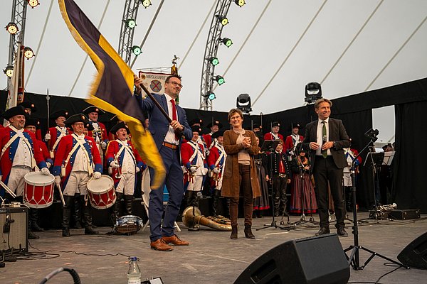 Zwei Männer und eine Frau auf der Bühne. Ein Mann hält eine Flagge in der Hand.
