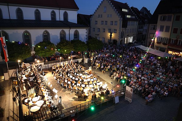 Ein großes Orchester spielt unter freiem Himmel bei Dämmerung vor einer großen Menschenmenge. Bunte Lichter schmücken die Umgebung, und historische Gebäude sind im Hintergrund sichtbar. Die Atmosphäre ist festlich und freundschaftlich.