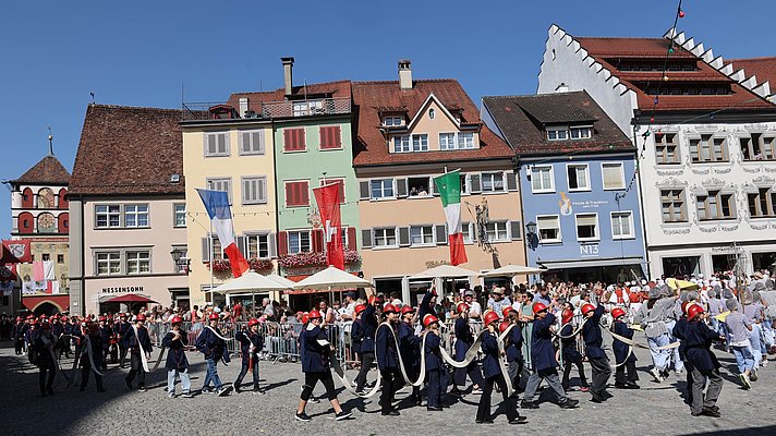 Kinder, die als Feuerwehrleute verkleidet sind und Schläuche dabei haben, gehen über den Marktplatz. Im Hingrund schauen viele Menschen zu. Die Häsuer strahlen in ihren bunten Farben, weil die Sonne darauffällt.