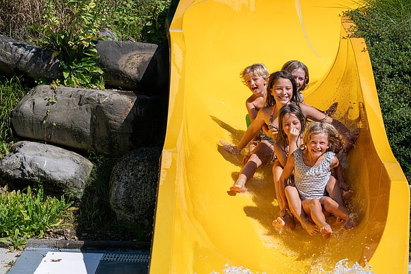 Fünf Kinder rutschen fröhlich gemeinsam eine große gelbe Wasserrutsche hinunter. Sie lachen und spritzen Wasser, während sie nach unten gleiten. Die Umgebung ist grün und sonnig, mit Steinen und Pflanzen im Hintergrund.