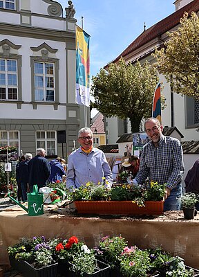 Zwei Männer lachen in die Kamera. Im Vordergrund Blumenkästen.