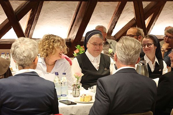 Schwester Bonavita sitzt umgeben von ihren Festgästen an einem festlich gedeckten Tisch. Alle lachen.