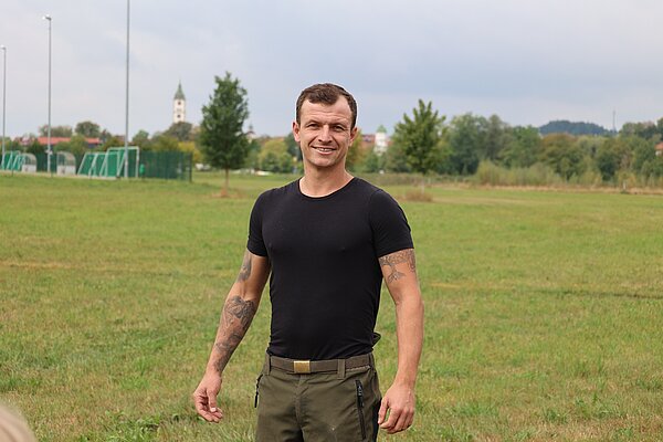 Ein Mann steht auf einem grünen Feld und lächelt. Er trägt ein schwarzes T-Shirt und grüne Hosen. Im Hintergrund sind Bäume und ein leicht bewölkter Himmel zu sehen. Einige Gebäude sind entfernt sichtbar.