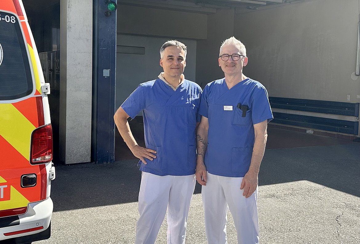 Zwei medizinische Fachkräfte in blauen Klinik-Outfits stehen lächelnd vor einem Krankenwagen in einer Garage. Im Hintergrund sind Teile der Garage und ein grünes Licht sichtbar.