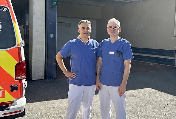 Zwei medizinische Fachkräfte in blauen Klinik-Outfits stehen lächelnd vor einem Krankenwagen in einer Garage. Im Hintergrund sind Teile der Garage und ein grünes Licht sichtbar.