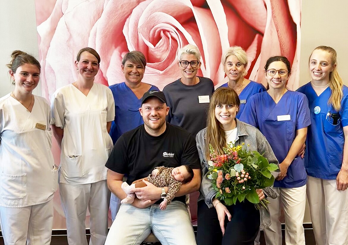 Eine Gruppe von neun Personen, darunter medizinisches Fachpersonal in Arbeitskleidung und ein Paar mit einem Neugeborenen sowie einem Blumenstrauß, posiert vor einer großen Wand mit Rosenmotiv.
