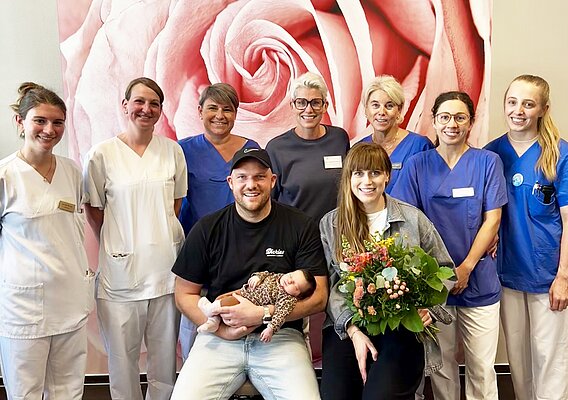 Eine Gruppe von neun Personen, darunter medizinisches Fachpersonal in Arbeitskleidung und ein Paar mit einem Neugeborenen sowie einem Blumenstrauß, posiert vor einer großen Wand mit Rosenmotiv.