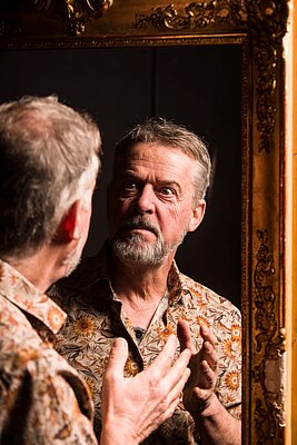 Ein Mann mit grauen Haaren und einem Bart betrachtet sein Spiegelbild in einem goldenen Rahmen. Er trägt ein blumiges Hemd und zeigt eine nachdenkliche, intensive Mimik, während er mit seinen Händen gestikuliert.