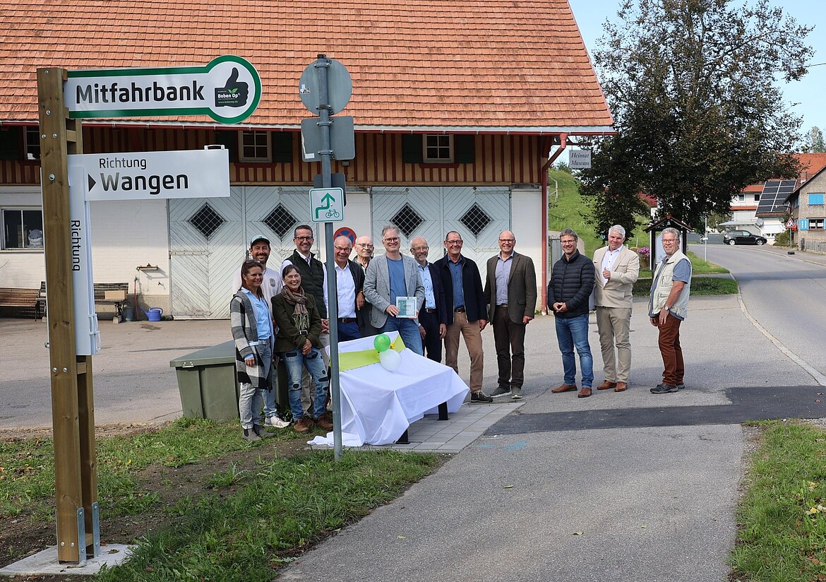 Eine Gruppe von Menschen steht an einem neuen Mitfahrbank-Schild in einem ländlichen Gebiet. Im Hintergrund sieht man ein rustikales Gebäude und eine Straße. Die Teilnehmer scheinen an einer Feier oder Einweihung teilzunehmen, einige lachen und posieren für ein Foto.