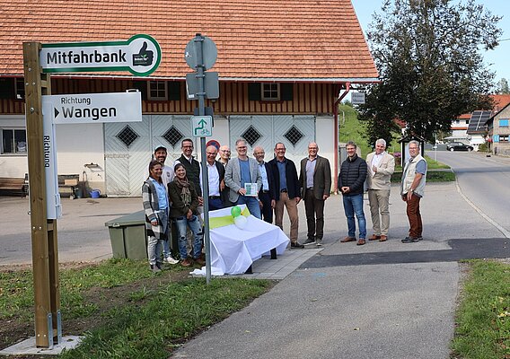 Eine Gruppe von Menschen steht an einem neuen Mitfahrbank-Schild in einem ländlichen Gebiet. Im Hintergrund sieht man ein rustikales Gebäude und eine Straße. Die Teilnehmer scheinen an einer Feier oder Einweihung teilzunehmen, einige lachen und posieren für ein Foto.