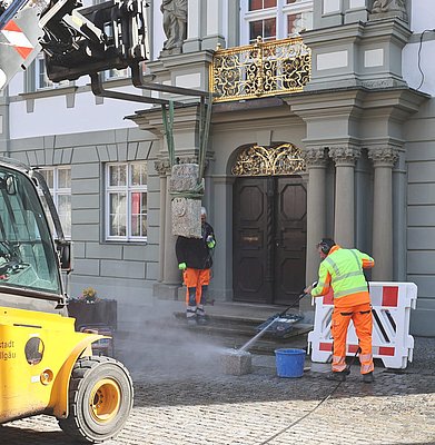 Ein Poller am Rathaus hängt an einem dicken Band am Kran. Der Sockel wird gereinigt.