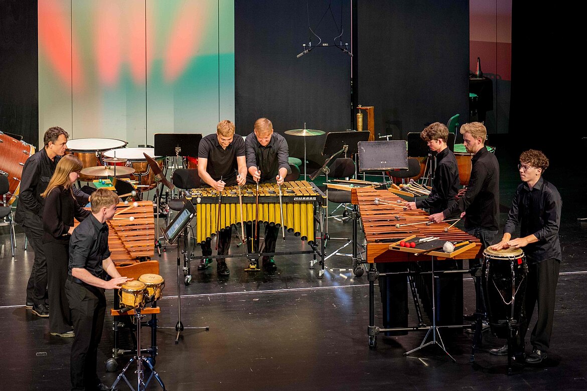 Eine Gruppe von fünf Musikern spielt auf verschiedenen Schlaginstrumenten, darunter Marimbas und Congas, auf einer Bühne. Sie tragen schwarze Kleidung und sind konzentriert bei der Aufführung. Im Hintergrund sind weitere musikalische Instrumente sichtbar.