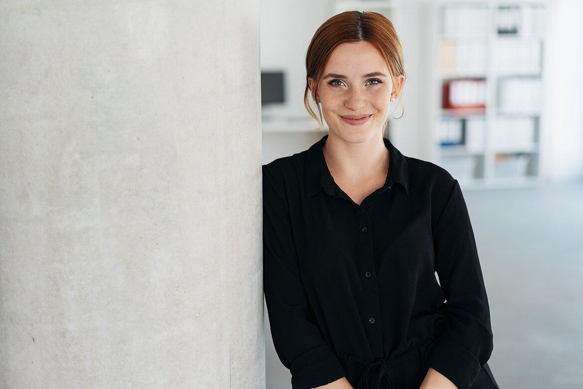Eine lächelnde Frau mit langen, roten Haaren steht an einem grauen Säule in einem modernen Büro. Sie trägt ein schwarzes Hemd und schaut freundlich in die Kamera. Im Hintergrund sind verschwommen Büroeinrichtungen zu sehen.
