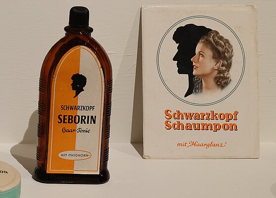 Eine braune Flasche mit schwarzem Deckel, beschriftet mit "Schwarzkopf Seborin Haar-Tonic", steht neben einer weißen Verpackung mit dem Titel "Schwarzkopf Schampo mit Haarglanz". Auf der Verpackung ist ein Profil einer Frau mit welligen Haaren abgebildet.