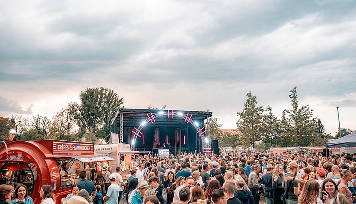 Ein großes Publikum versammelt sich vor einer Bühne im Freien, die von bunten Lichtern beleuchtet wird. Im Vordergrund sind Food-Trucks sichtbar. Der Himmel ist bewölkt und es herrscht eine lebhafte Atmosphäre auf dem Event.