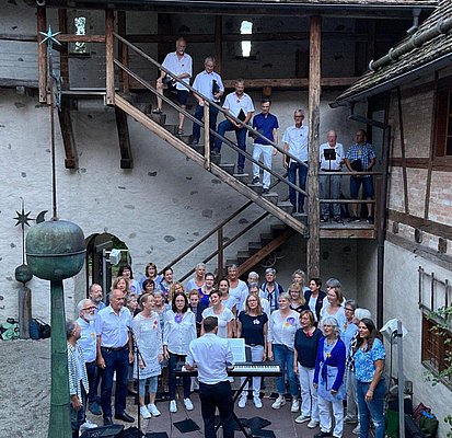 Eine Gruppe von Sängern steht in einem Innenhof vor einer Treppe, auf der einige Mitglieder stehen. Ein Dirigent mit einem Keyboard leitet den Chor. Die Teilnehmer tragen überwiegend helle Kleidung und einige halten Notenblätter. Die Umgebung hat historische Architektur.