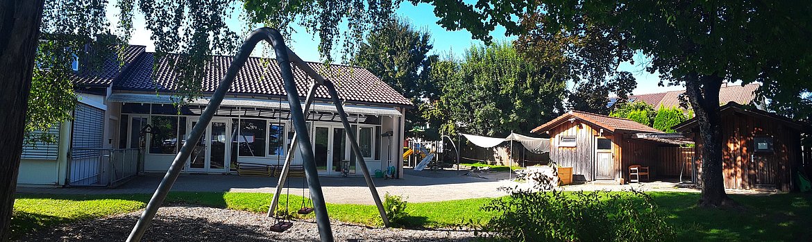 Eine kindersichere Außenanlage mit einem Spielplatz wird von einem modernen Gebäude und einem Holzhaus umgeben. Die Umgebung ist grün und sonnig, mit einer Schaukel im Vordergrund und einem gepflasterten Bereich zum Spielen.