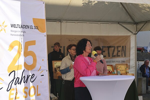 Eine Veranstaltung im Freien mit einer Frau, die ein Mikrofon hält und spricht. Im Hintergrund sind Personen zu sehen, die aufmerksam zuhören. Ein großes Banner mit der Aufschrift „25 Jahre EL SOL“ ist im Vordergrund.