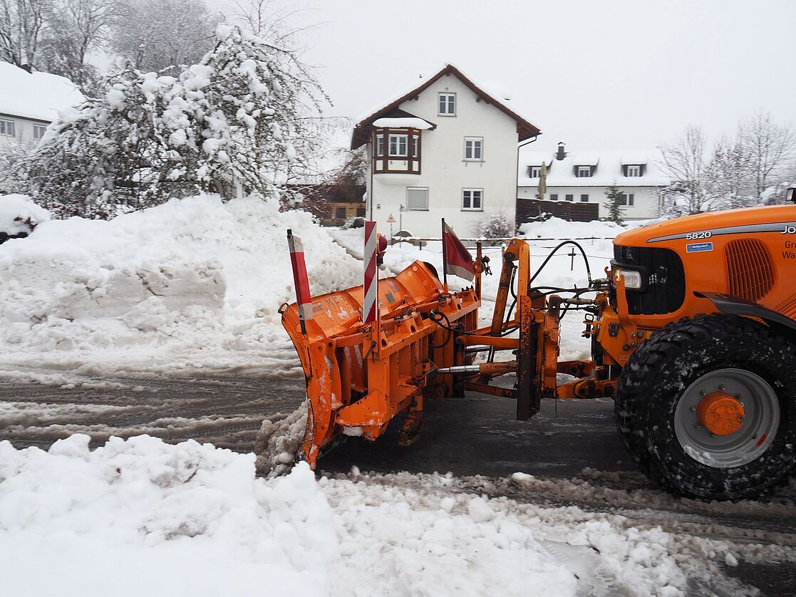 Ein orangefarbener Schneepflug steht in einer verschneiten Straße. Im Hintergrund sind schneebedeckte Bäume und Häuser zu sehen. Der Pflug ist bereit, Schneemassen von der Straße zu räumen.