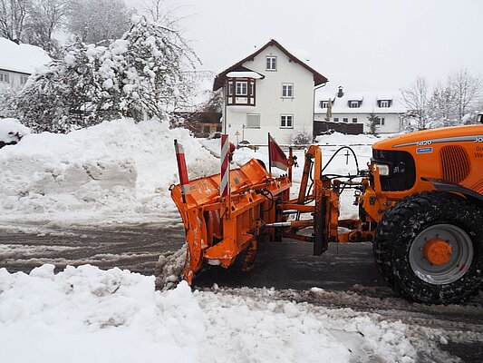 Ein orangefarbener Schneepflug steht in einer verschneiten Straße. Im Hintergrund sind schneebedeckte Bäume und Häuser zu sehen. Der Pflug ist bereit, Schneemassen von der Straße zu räumen.