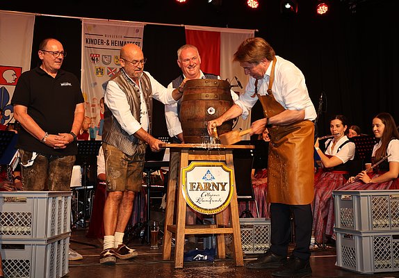 Eine Gruppe von Männern in traditioneller Kleidung steht auf einer Bühne. Einer von ihnen zapft aus einem großen Holzfass. Im Hintergrund sitzen Personen in Trachten und das Ambiente ist festlich dekoriert.