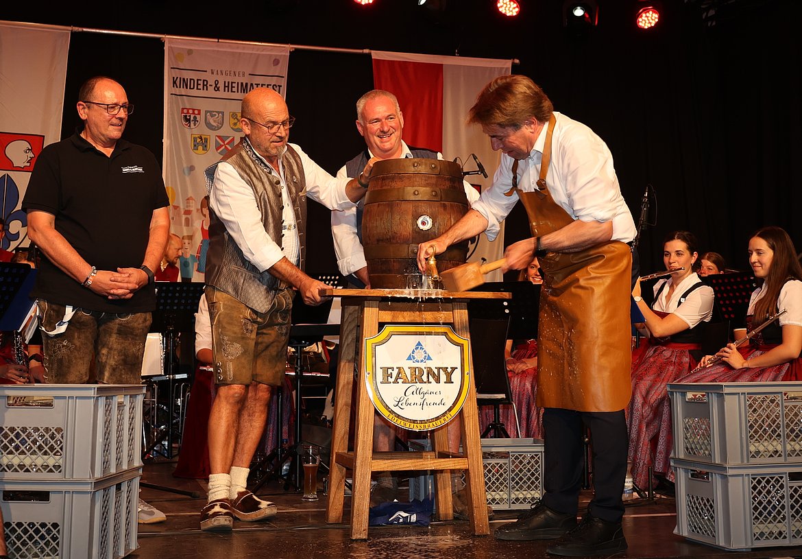 Eine Gruppe von Männern in traditioneller Kleidung steht auf einer Bühne. Einer von ihnen zapft aus einem großen Holzfass. Im Hintergrund sitzen Personen in Trachten und das Ambiente ist festlich dekoriert.
