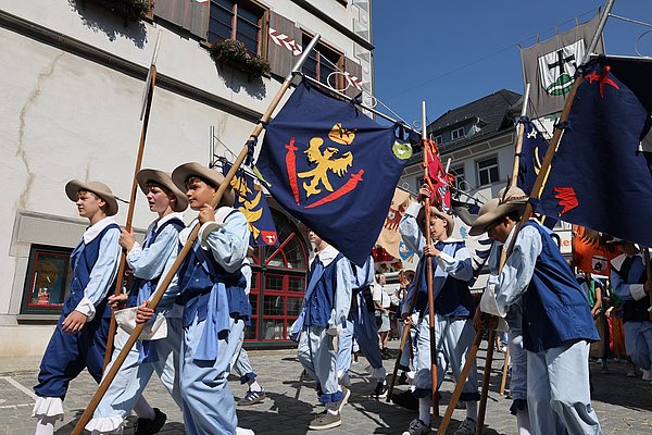 Eine Gruppe von Kindern in historischen Kostümen marschiert durch die Straße. Sie tragen Fahnen mit Wappen und Symbolen. Die Szene spielt sich an einem sonnigen Tag ab, umgeben von Gebäuden.