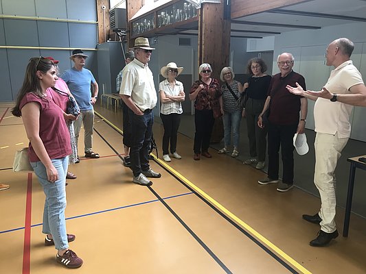 Eine Gruppe von Personen steht in einer Halle. Die Leute sind unterschiedlichen Alters und tragen Freizeitkleidung. Einige lachen und sprechen miteinander, während eine Person am rechten Bildrand spricht und gestikuliert. Die Atmosphäre wirkt informell und freundlich.