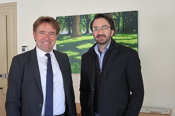Zwei Männer stehen lächelnd in einem Raum. Der Mann links trägt einen Anzug und eine Krawatte, der Mann rechts hat eine Brille und einen dunklen, gestreiften Blazer an. Im Hintergrund ist ein Bild mit einer grünen, lichten Waldlandschaft zu sehen.