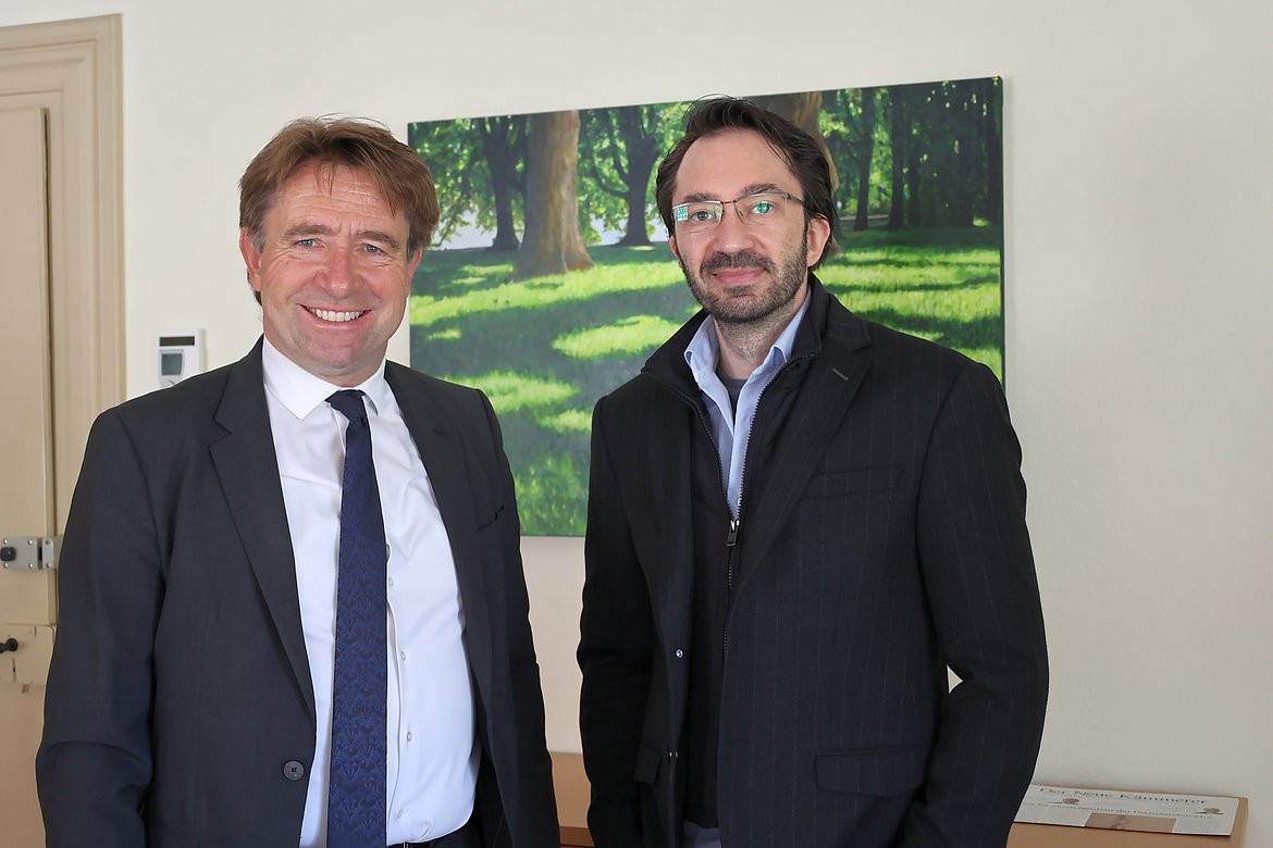 Zwei Männer stehen lächelnd in einem Raum. Der Mann links trägt einen Anzug und eine Krawatte, der Mann rechts hat eine Brille und einen dunklen, gestreiften Blazer an. Im Hintergrund ist ein Bild mit einer grünen, lichten Waldlandschaft zu sehen.