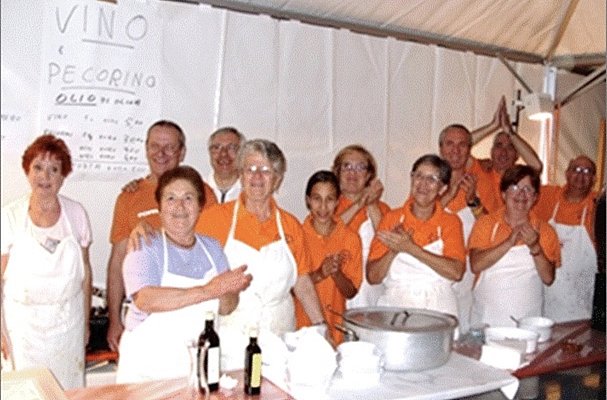 Eine Gruppe von mehreren Personen in orangefarbenen T-Shirts und Schürzen steht in einem Zelt. Sie lächeln und applaudieren, während sie sich um einen Tisch mit Kochutensilien und einer großen Schüssel versammeln. Im Hintergrund ist ein Schild mit Speisen und Getränken zu sehen.