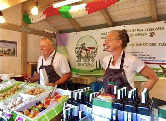 Zwei Personen stehen an einem Verkaufsstand in einem Holzgebäude. Sie lächeln und präsentieren eine Auswahl an frischen Lebensmitteln, darunter Obst, Gemüse und Öle. Im Hintergrund hängt ein Banner mit der Aufschrift "Agriturismo Biancospino".