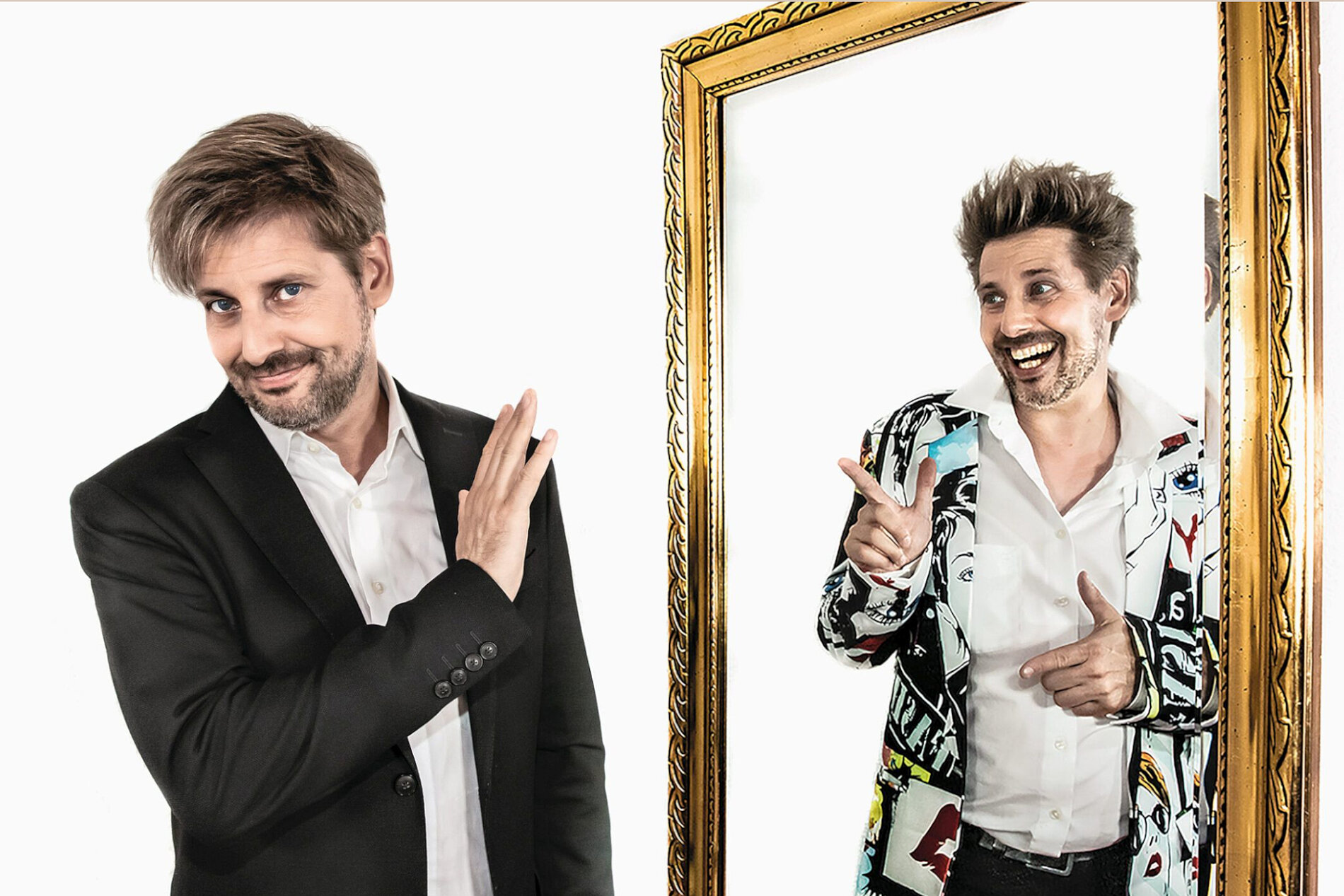 Ein Mann in einem schwarzen Anzug steht auf der linken Seite und winkt mit der Hand. Auf der rechten Seite spiegelt ihn ein Bild in einem goldenen Rahmen, in dem er einen bunten, gemusterten Anzug trägt und fröhlich lächelt, während er mit den Händen gestikuliert.