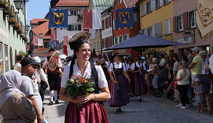 Eine Frau in einem dirndl geht mit Blumen in der Hand vor einer Musikapelle. Am Straßenrand stehen Menschen und schauen zu. Die Straße ist geschmückt.
