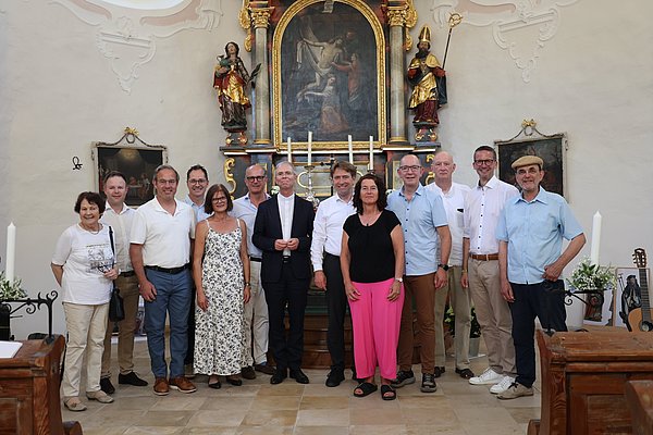Gruppenfoto von 15 Personen in einer Kirche. Sie stehen vor einem Altar mit religiösen Figuren und Gemälden. Die Anwesenden tragen verschiedene, ggf. sommerliche Kleidung und lächeln in die Kamera. Die Kirche hat eine helle, einladende Atmosphäre.
