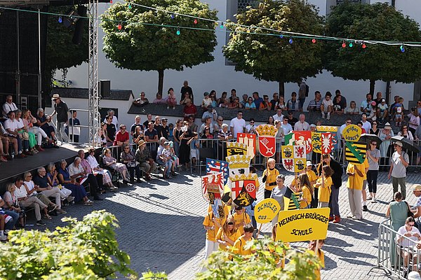 Eine Menschenmenge verfolgt eine festliche Veranstaltung auf einem Platz. Schüler in gelben T-Shirts tragen bunte Schilder mit Wappen und Aufschriften. Einige Zuschauer sitzen auf einer Tribüne, während andere auf Steinbänken stehen. Es ist sonnig und eine festliche Stimmung herrscht.