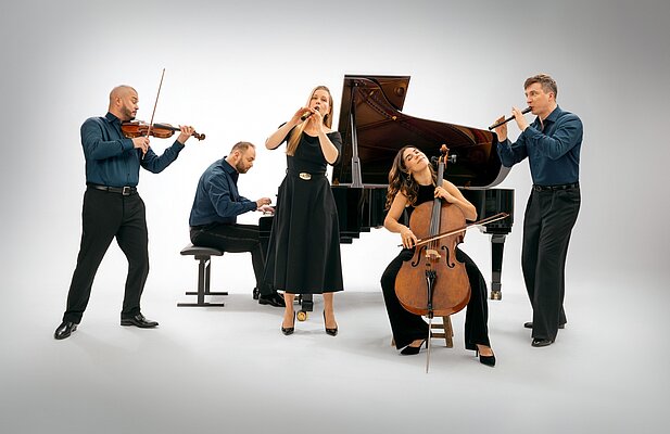 Eine Gruppe von fünf Musikern steht um ein Klavier. Ein Violinist und ein Flötist spielen, während eine Cellistin und eine Sängerin auftreten. Ein Pianist begleitet sie am Klavier. Alle tragen formelle Kleidung und zeigen eine lebendige Bühnendarstellung.