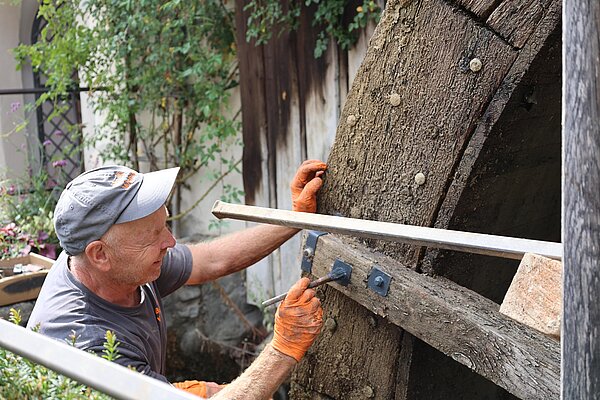 Ein Handwerker arbeitet an einem großen hölzernen Rad, das Teil einer Mühle oder ähnlichen Struktur ist. Er trägt Handschuhe und hat eine Werkzeugspritze in der Hand. Im Hintergrund sind Pflanzen und eine Wand sichtbar.