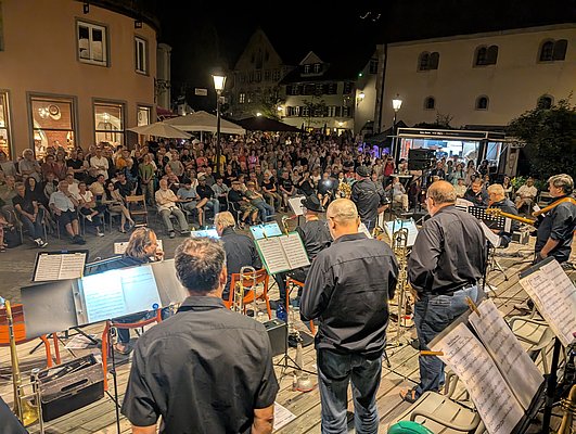 Eine Band spielt auf einer Bühne vor einer großen Menschenmenge in einer Stadt. Die Musiker sind in schwarzen Hemden gekleidet, während das Publikum auf Stühlen sitzt und dem Auftritt aufmerksam folgt. Es ist Nacht, beleuchtet von Straßenlaternen und Bühnenlichtern.