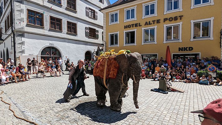Eine Figur, die aussieht wie ein Elefant steht auf einem Platz, während ein Mensch ihn an der Leine führt. Viele Zuschauer sitzen und stehen um den Platz herum. Im Hintergrund sind Gebäude und ein Schild des Hotels "Alte Post" zu sehen.