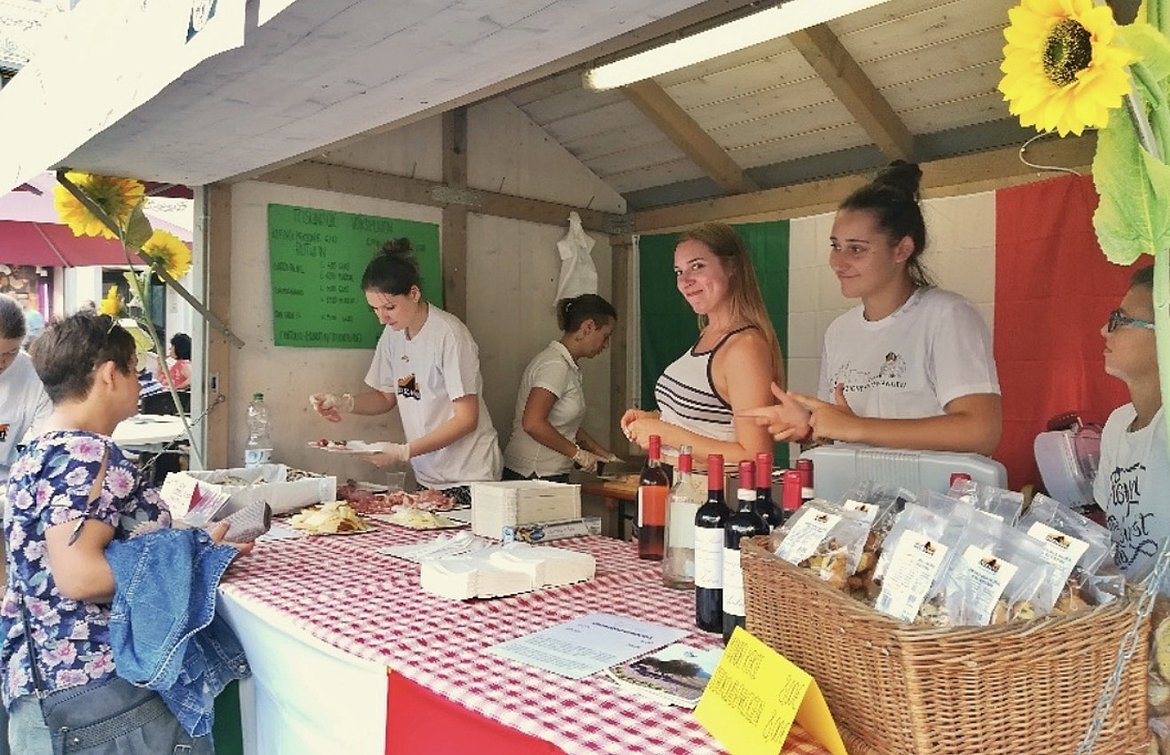 Auf einem Marktstand stehen mehrere Verkäuferinnen hinter einer Theke mit Lebensmitteln und Getränken. Die Theke ist mit rotem und grünem Tuch dekoriert. Im Vordergrund sind interessierte Käufer zu sehen, die sich um den Stand versammeln.