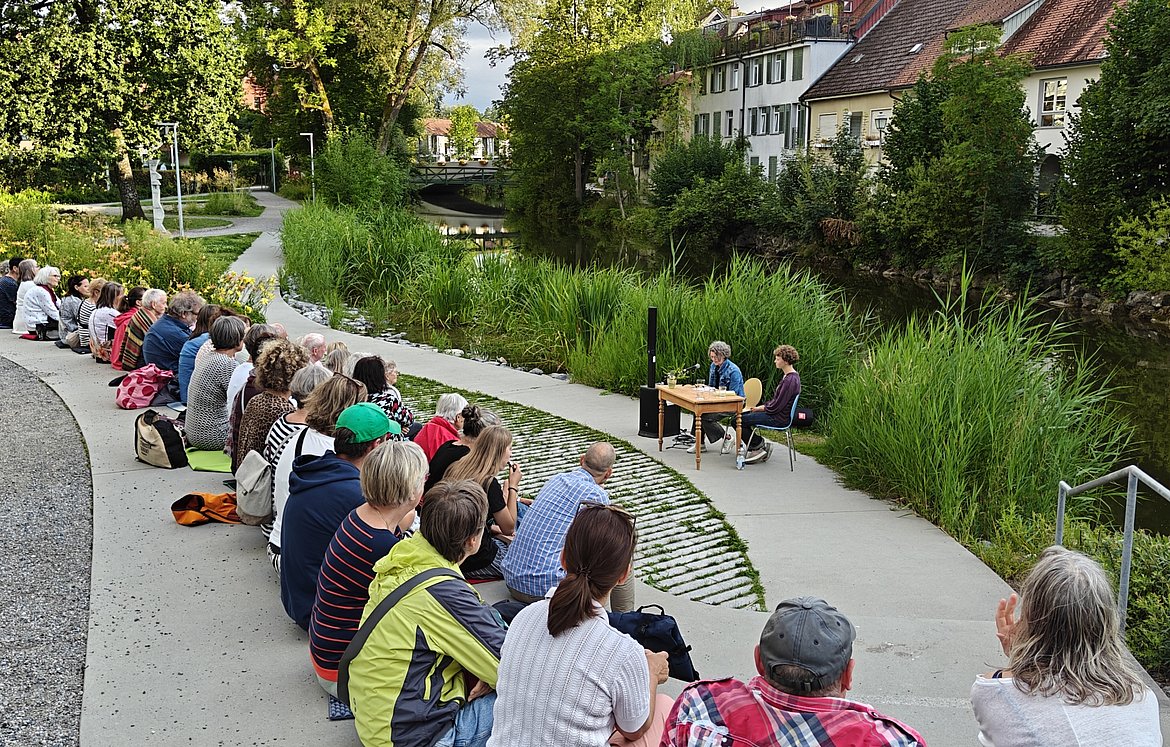 Eine Gruppe von Menschen sitzt auf Stufen entlang der Argen in Wangen. Im Hintergrund sind zwei Personen zu sehen, die an einem Tisch sitzen und sprechen. Grüne Pflanzen umgeben den Bereich, und Häuser der Altstadt sind in der Ferne sichtbar.