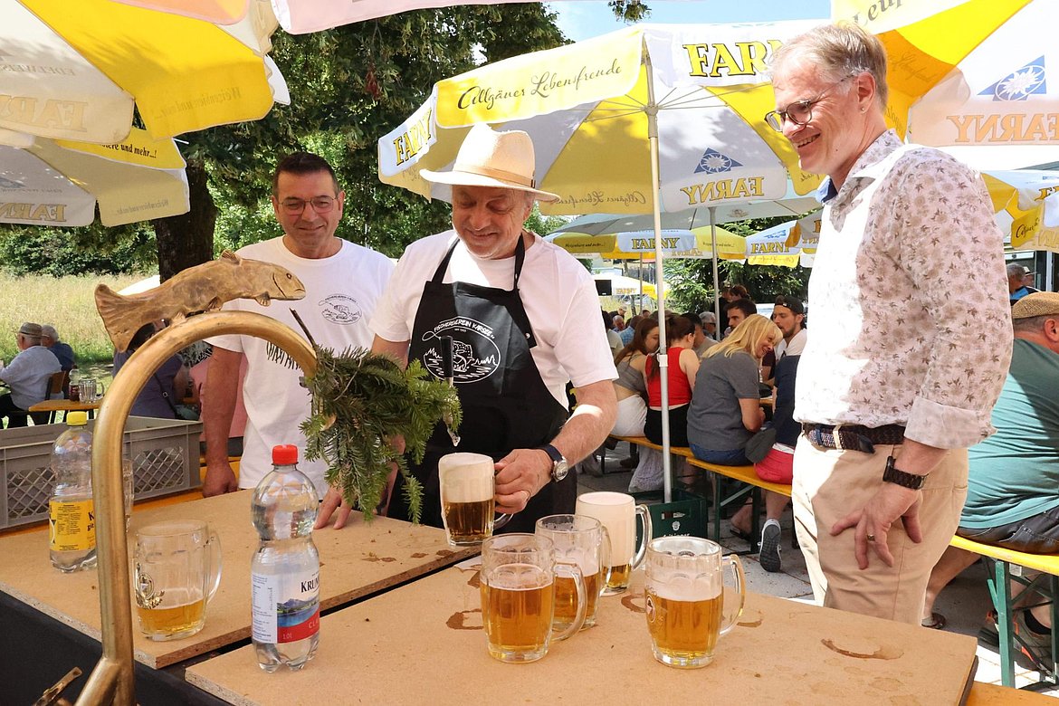 Drei Männer stehen an einem Brunnen, aus dem man Bier zapfen kann.